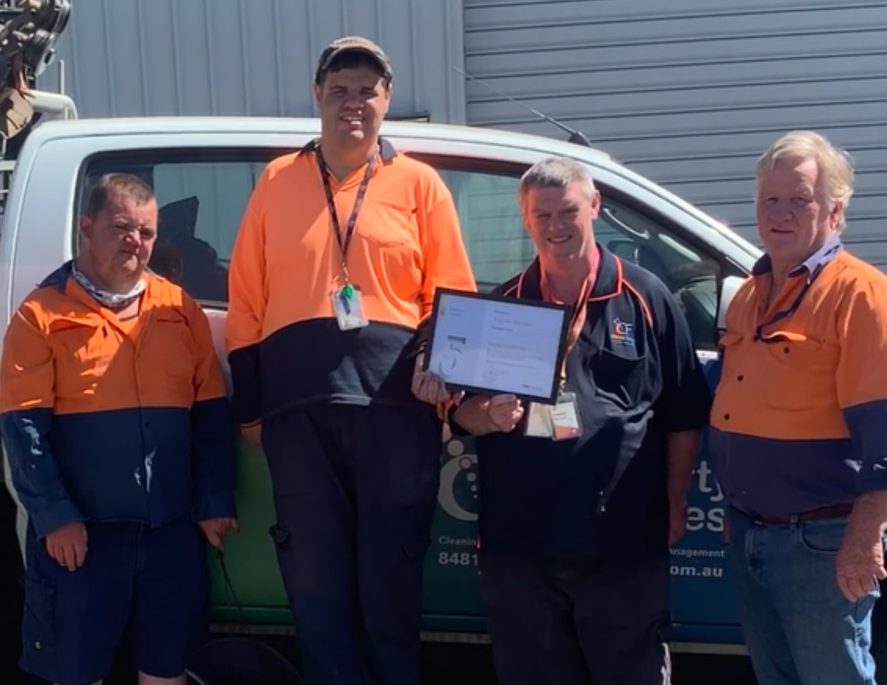 Four men standing in front of a van. One man is holding a certificate.